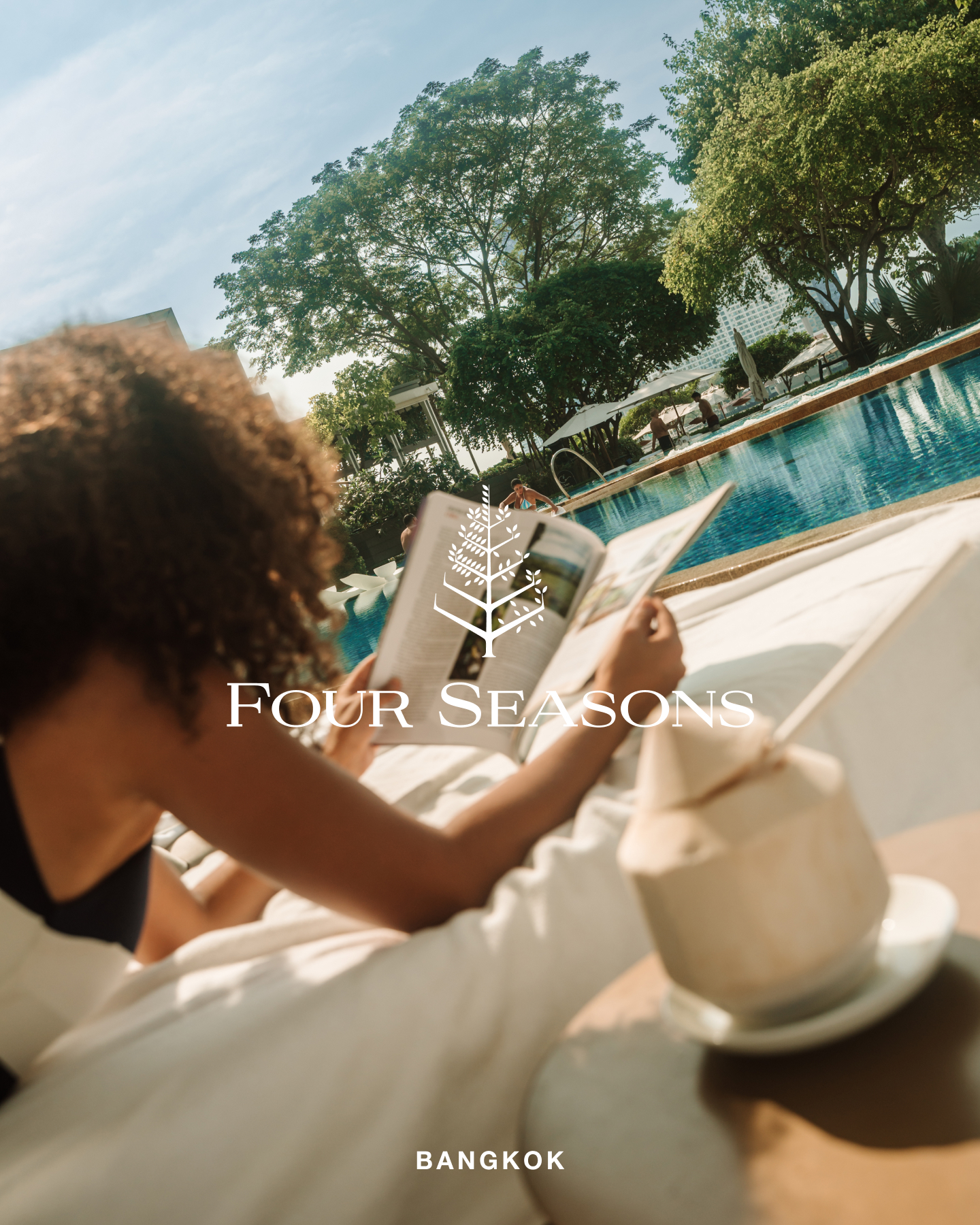 A woman with curly hair is relaxing on a lounge chair by a swimming pool, reading a magazine. She is holding a coconut drink. The Four Seasons logo and name are overlaid on the image, along with the text "BANGKOK" at the bottom. The scene is bright and sunny, with lush green trees and blue water visible. The overall impression is one of luxury and relaxation.