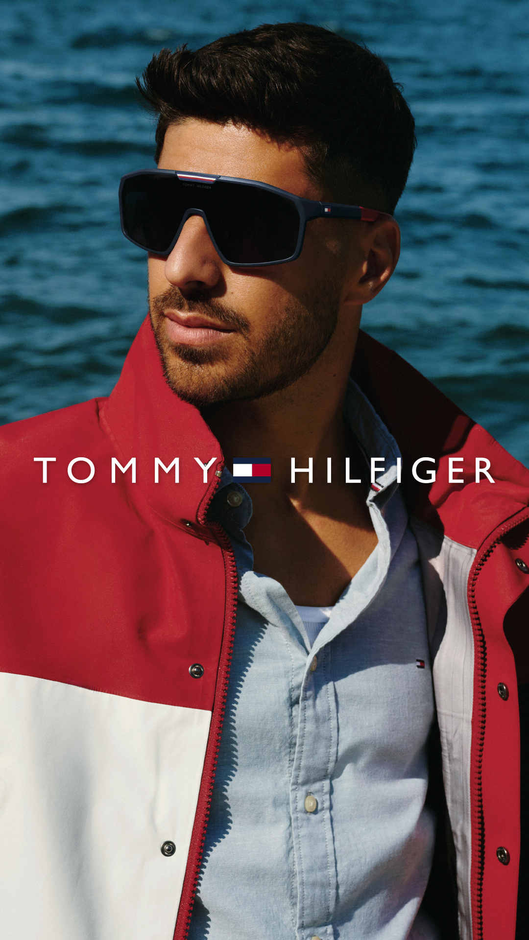 A man wearing sunglasses and a red and white jacket over a light blue button-down shirt stands against a backdrop of blue water. The Tommy Hilfiger logo and name are displayed prominently across the man's chest. The image is a close-up, focusing on the man's upper body and the branding. There is no explicit offer or call to action within the visual creative itself.