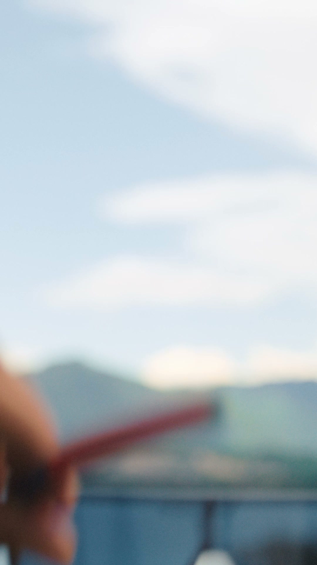 The image is blurred, showing a hand holding a red object in the foreground against a backdrop of hazy mountains and a light blue sky with clouds. Text overlays the image, announcing the discovery of the new Tommy Hilfiger Eyewear collection and tagging the brand's account with a hashtag.