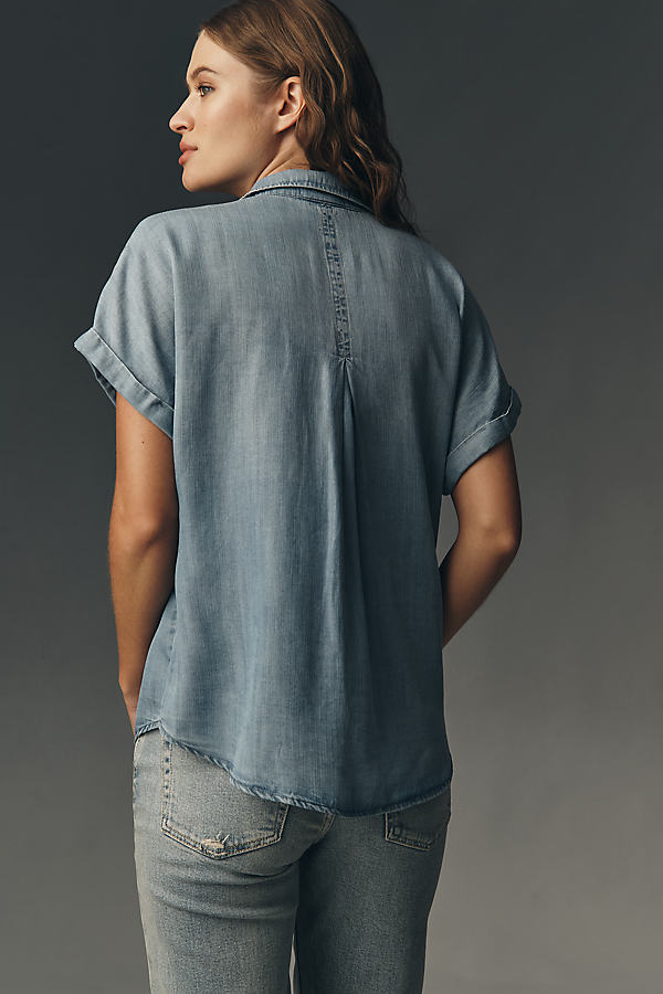 A woman is shown from the back, wearing a light blue denim shirt with cuffed short sleeves and light-wash jeans. The shirt features a subtle pleat down the center back and a collar. The background is a solid dark gray. The text "Give a warm welcome to new tops." is overlaid on the image.