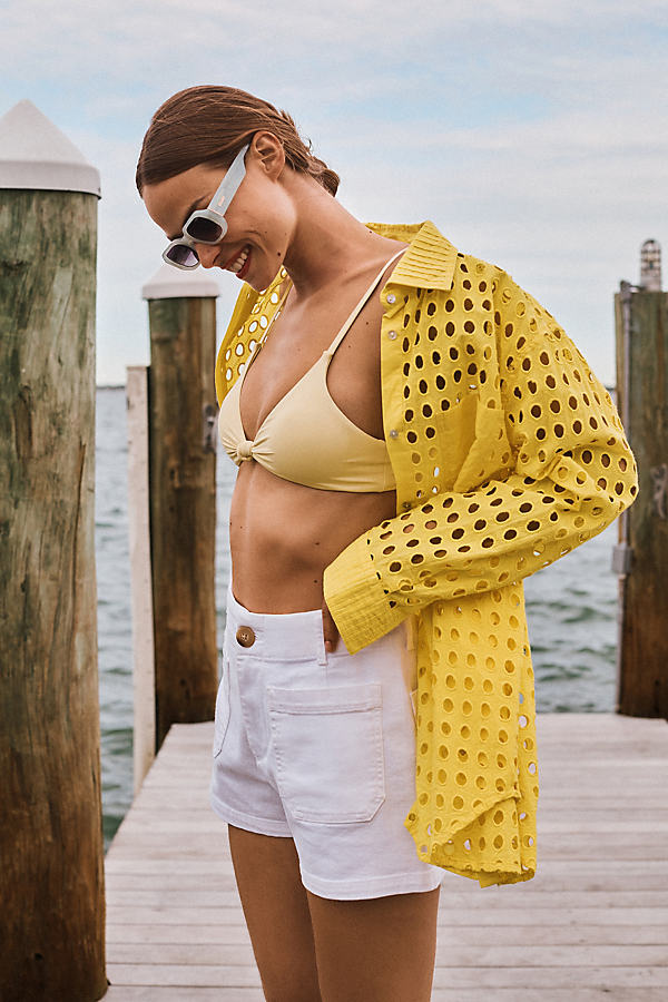 A woman wearing a yellow eyelet shirt open over a yellow bikini top and white shorts stands on a wooden pier. She is looking down and smiling, with her hand in her pocket. White sunglasses are on her head. The background shows a body of water and wooden posts. The text "These pieces = easiest outfit repeaters." is overlaid on the image.