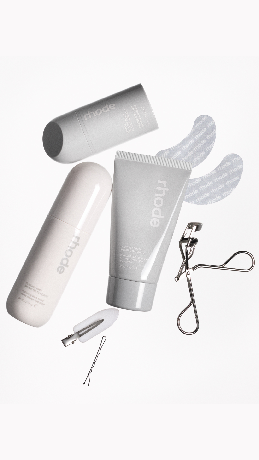 A flat lay of Rhode skincare products is arranged against a white background. The products include a spray bottle, a tube, and a smaller bottle, along with an eyelash curler and hairpins. The text "get ready with rhode 🤍" is overlaid on the image. The products appear to be in motion or floating, creating a dynamic composition.