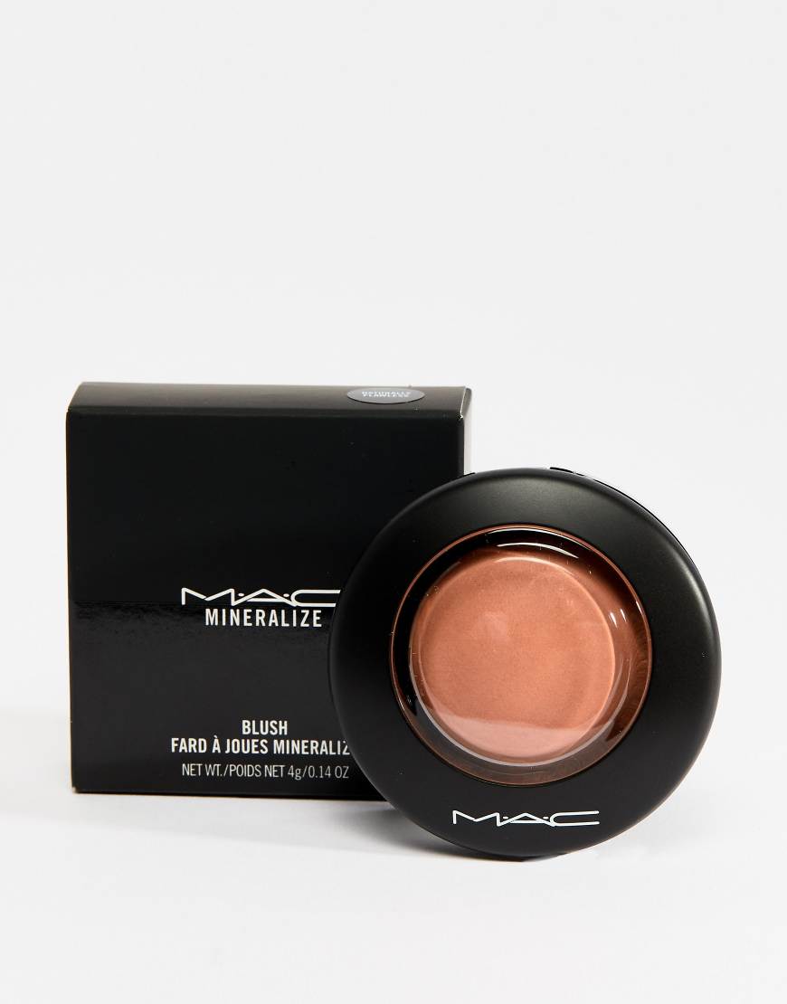 A MAC Mineralize blush compact and its black packaging are displayed against a white background. The blush compact is open, revealing a peachy-pink powder. The packaging has the MAC logo and product details printed on it. There is no text overlay on the image itself.