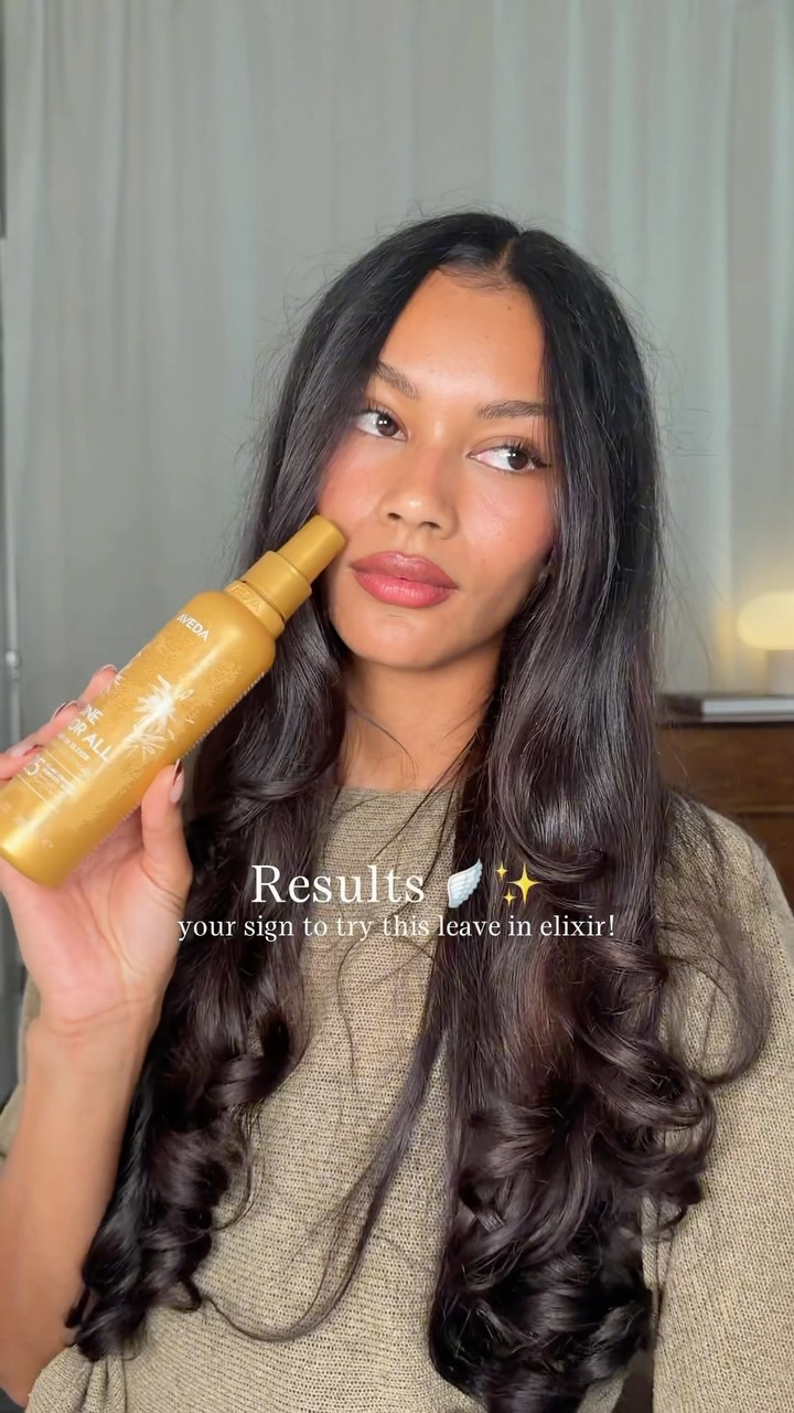 A woman with long, dark, wavy hair holds a gold bottle of Aveda leave-in elixir. She is looking off to the side with a slight smile. Text overlays the image, reading "Results" with a small wing icon and sparkles, followed by "your sign to try this leave in elixir!". The ad copy mentions the product is the NEW Aveda One For All Leave-In Elixir and is an #AvedaPartner.