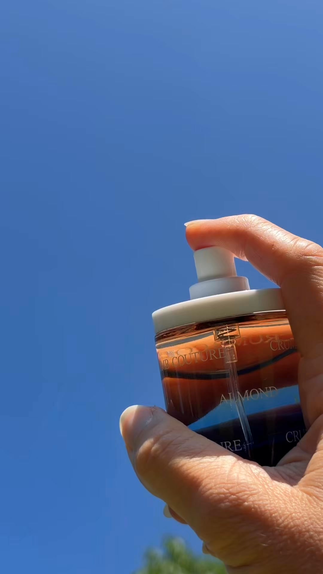 A hand holds a clear glass perfume bottle with a white spray top against a bright blue sky. The bottle contains a warm, amber-colored liquid. Text overlays the image, with the main headline in a large, bold font. The copy describes the scent of the perfume, highlighting notes of almond cream, powdered sugar, and buttery musks, suggesting it will be appealing and mouth-watering.