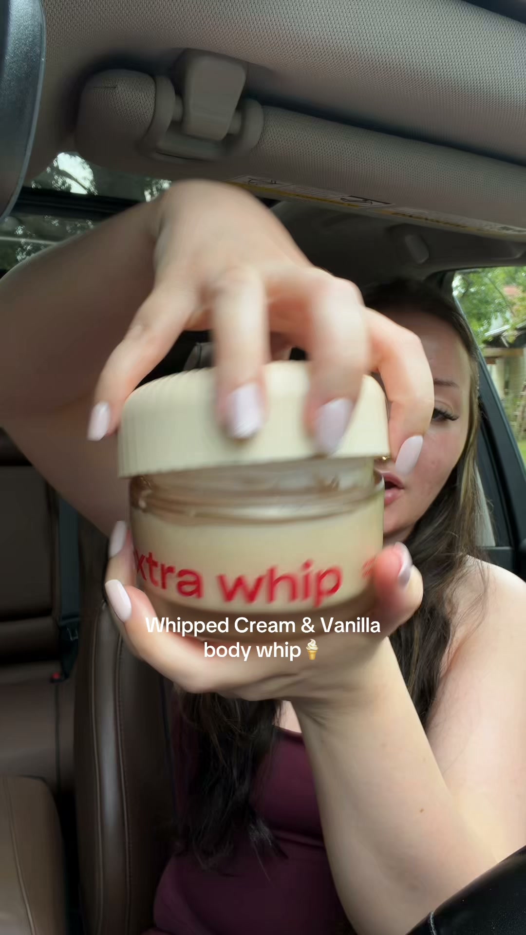 A person is holding a jar of "Extra Whip" body whip in a car. The jar has red text that says "Extra Whip" and below that, white text reads "Whipped Cream & Vanilla body whip" with an ice cream cone emoji. The person's hands are visible, holding the jar up to the camera. The background shows the interior of a car and a blurred outdoor scene.