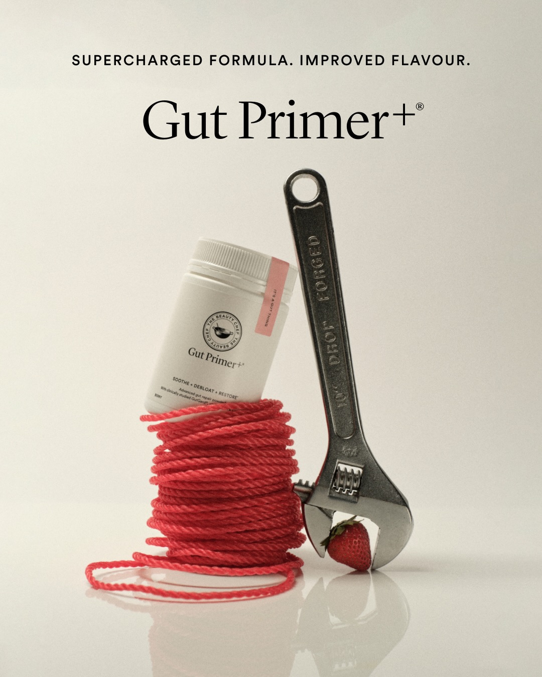 A white bottle of "Gut Primer+" is wrapped in a coil of red rope. A metal wrench is positioned to grip a single strawberry, with the wrench's jaws open around it. The text "SUPERCHARGED FORMULA. IMPROVED FLAVOUR." is displayed prominently at the top of the image, above the product name "Gut Primer+".