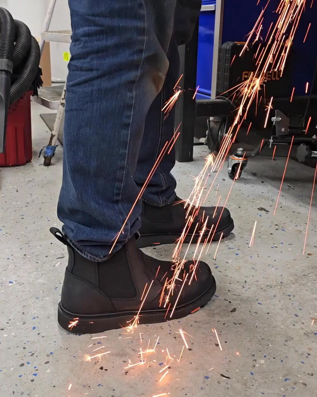 A close-up shot focuses on a person's feet wearing black work boots, with sparks flying around them, suggesting a work environment. The boots are a Chelsea boot style with elastic side panels and a pull tab at the back. The text overlay reads "Pull on the Ohman and stay comfortable all day. Breathable. Cushioned. Free shipping." The image highlights the durability and comfort of the work boots in a demanding setting.
