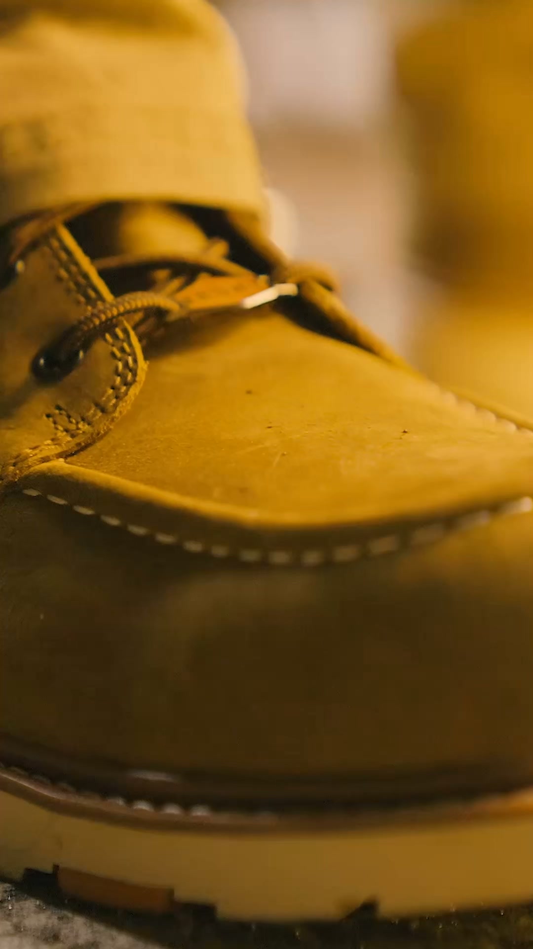 A close-up, shallow depth-of-field shot focuses on the toe and laces of a tan work boot. The stitching and texture of the leather are visible. The background is blurred and out of focus, with a warm, golden hue. Text overlays the image, stating 'You asked, we listened. Now you can find BRUNT at Academy Sports + Outdoors. Click 'Learn More' to find a location near you.'