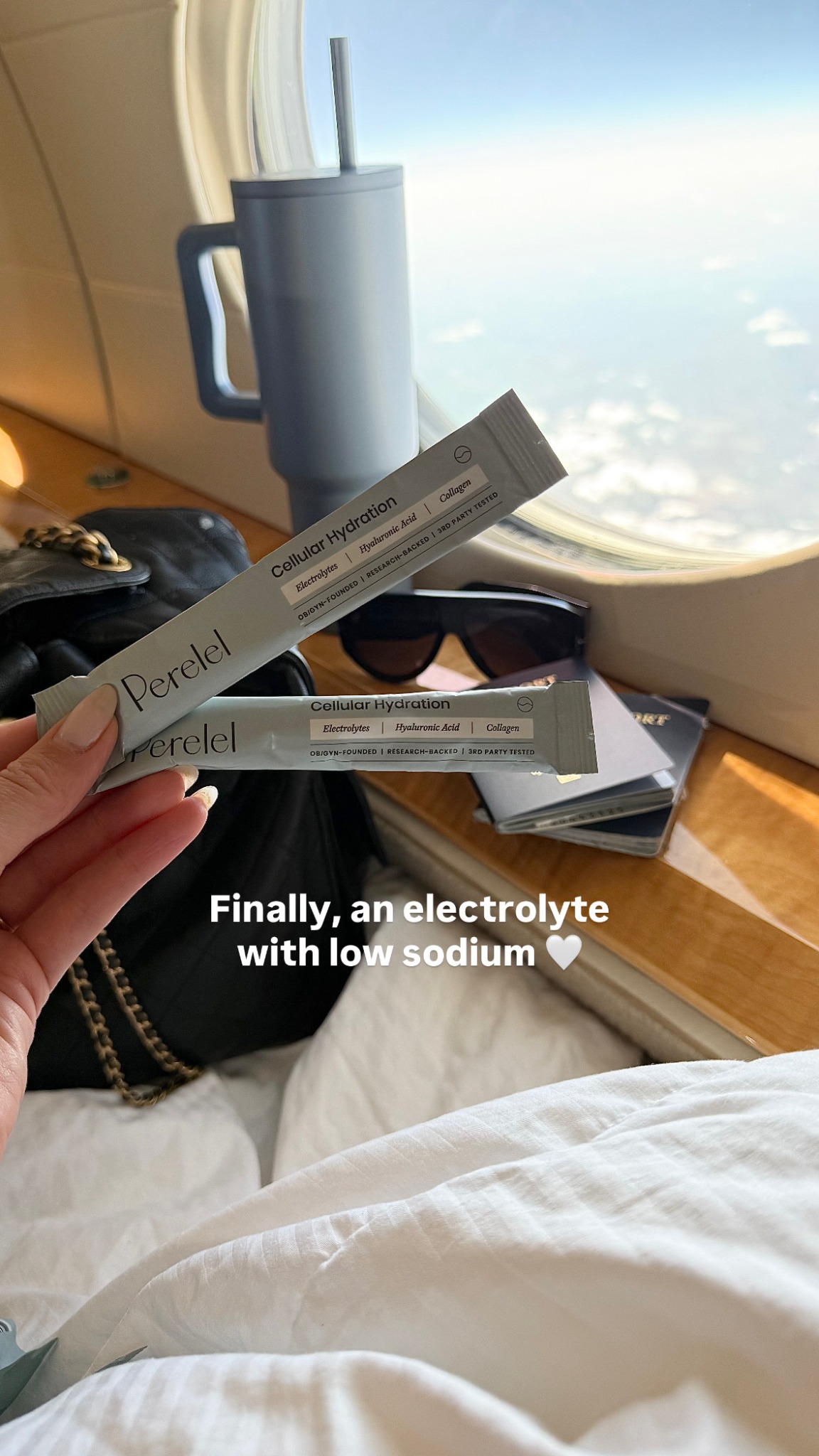 The image shows two Perelel Cellular Hydration powder packets held by a person's hand, with a blue travel mug and sunglasses in the background. The scene appears to be inside a private jet, with the sky visible through the window. On-screen text reads "Finally, an electrolyte with low sodium " followed by a white heart emoji. The copy provides context about the product's benefits, including low sodium, hyaluronic acid, and collagen.