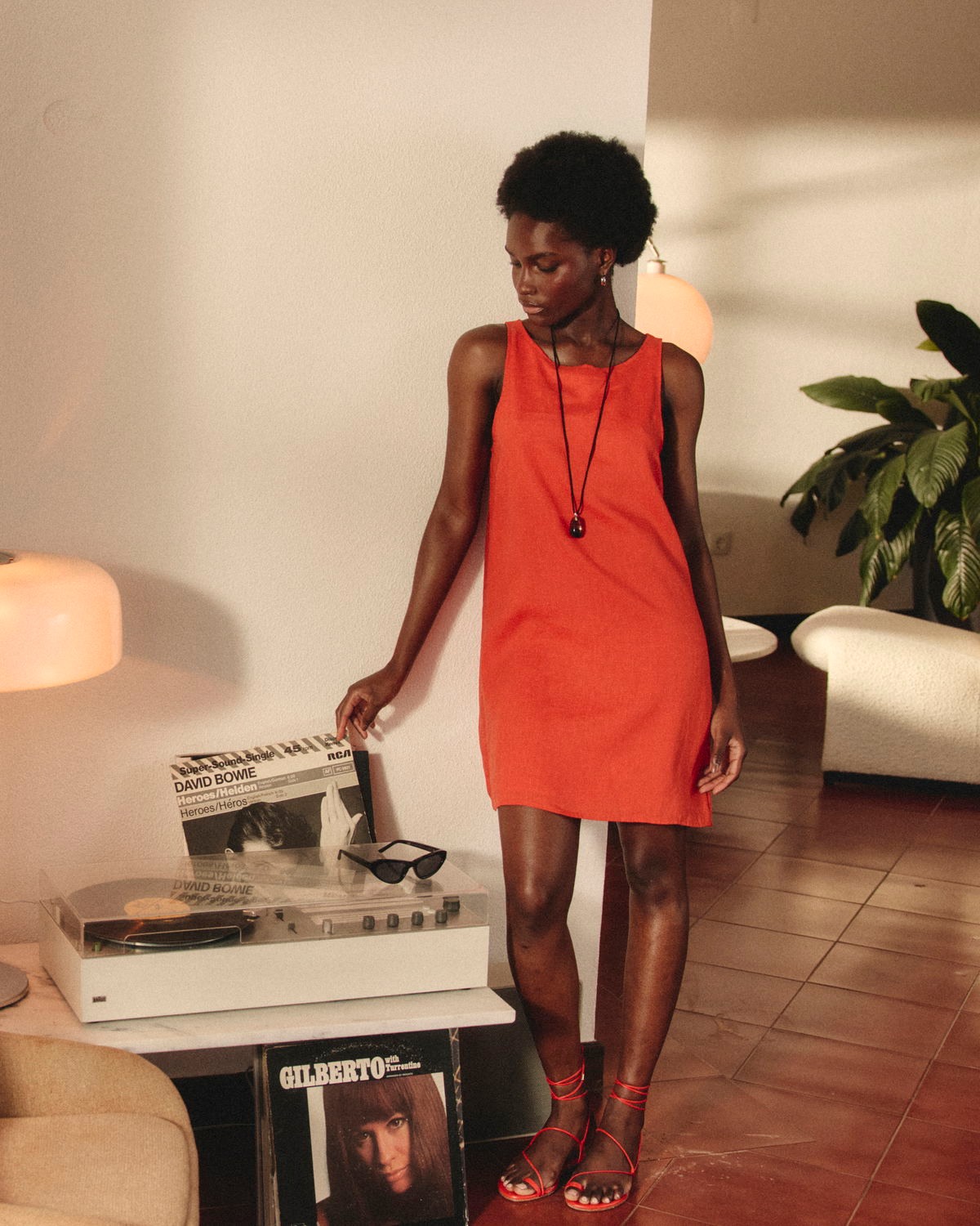 A young Black woman stands in a well-lit room, wearing an orange sleeveless dress and red strappy sandals. She is looking down at a vintage record player on a shelf, with her hand resting on it. Sunglasses are placed on the record player. The text "100% lin français Matière ultra respirante et légère Doublure en Ecovero, 0 transparence" is overlaid on the image. The scene has a retro aesthetic with warm lighting and plants in the background.