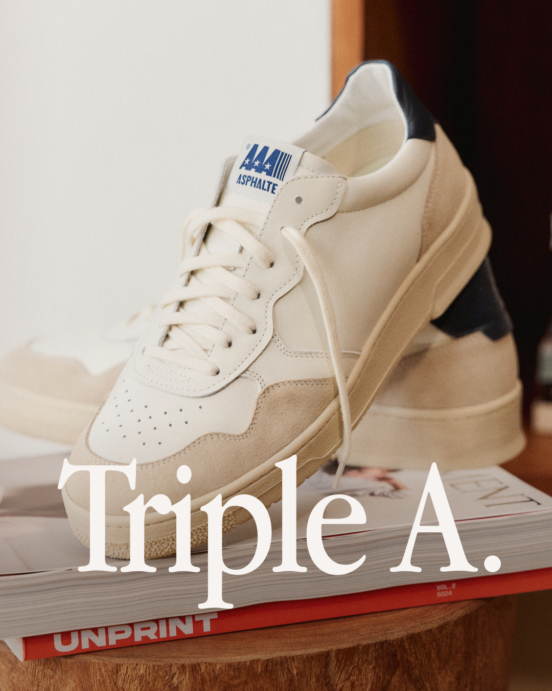 A close-up, eye-level shot focuses on a pair of cream-colored sneakers with beige suede accents and navy blue heel accents. The sneakers are placed on top of a stack of magazines, with the word "Triple A." prominently displayed in a large, serif font across the front of the shoes and magazines. The background is softly blurred, keeping the focus on the product and the text overlay. The text overlay reads "Oh, ça ? C’est juste une excellente paire de baskets." which translates to "Oh, this? It's just an excellent pair of sneakers."