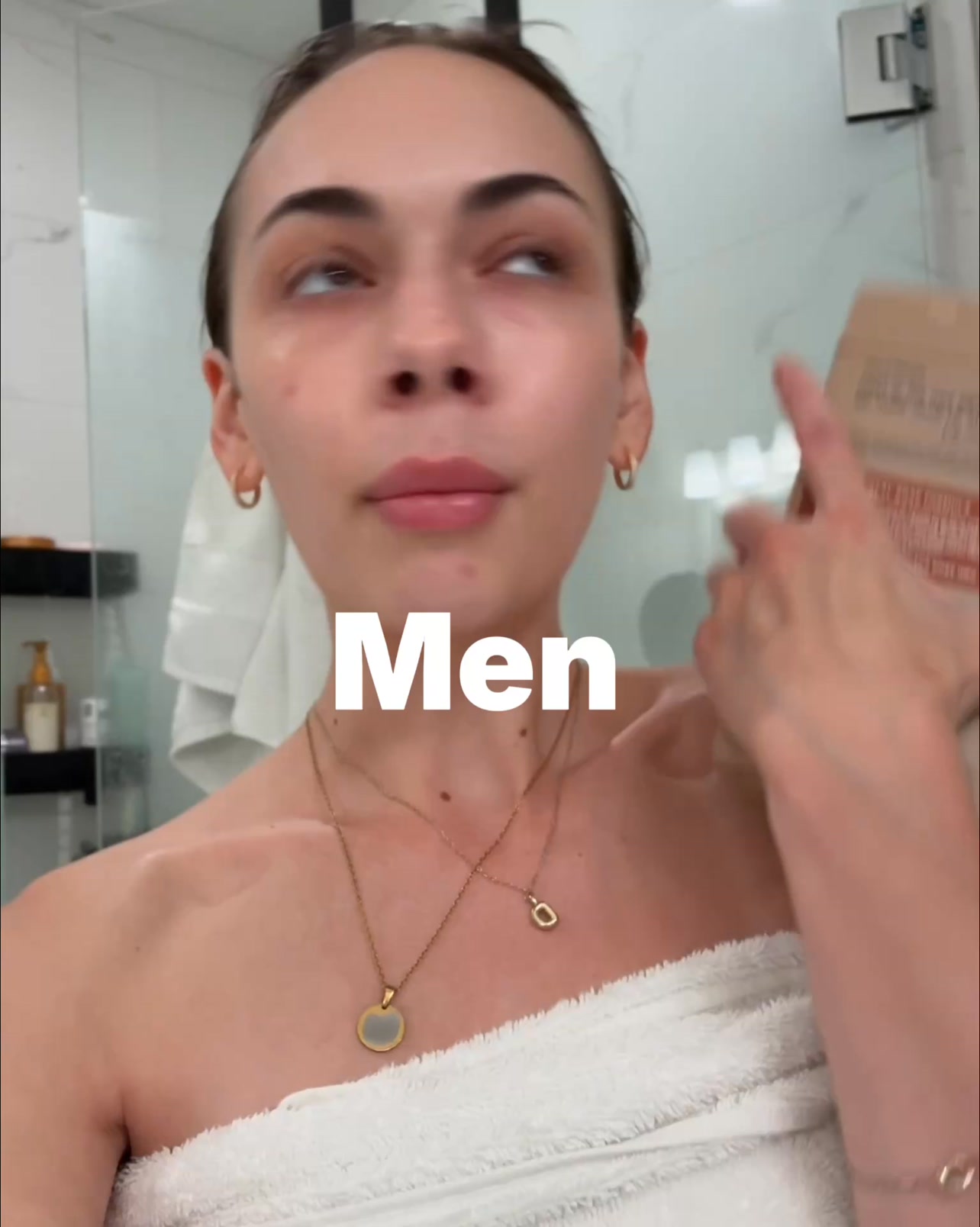 A woman is shown from the chest up, wrapped in a white towel, with two gold necklaces. She is looking upwards and to the side. Large white text overlays the image, reading "Men". The background appears to be a bathroom with a glass shower door. The woman is holding a small brown box in her right hand near her face.