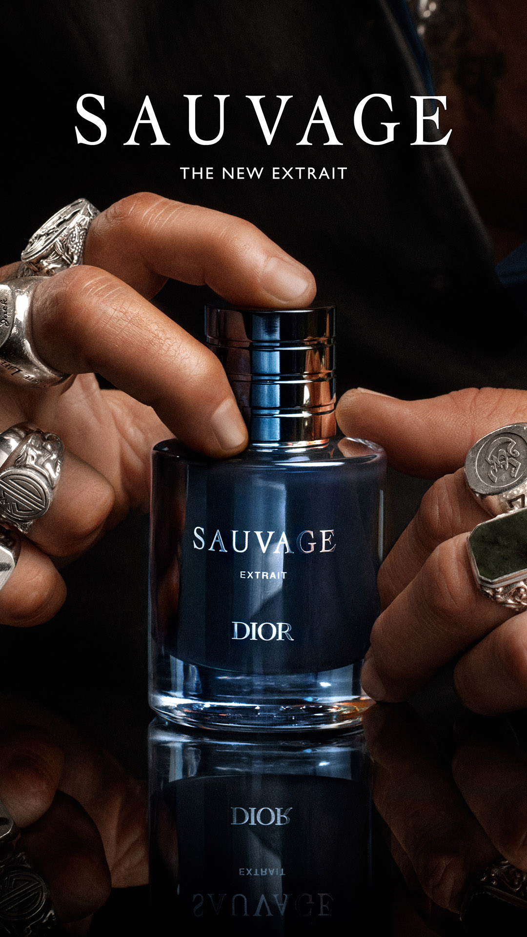 A close-up shot focuses on a man's hands, adorned with ornate rings, holding a dark blue glass bottle of Dior Sauvage Extrait perfume. The bottle is sleek and modern, with a metallic blue and silver cap. The background is dark and slightly blurred, emphasizing the product. Text overlays on the image read "SAUVAGE, DAS NEUE EXTRAIT" at the top and "DIOR" near the bottom, with the product name and "EXTRAIT" also visible on the bottle itself. The overall aesthetic is luxurious and sophisticated, highlighting the fragrance and its association with Johnny Depp.