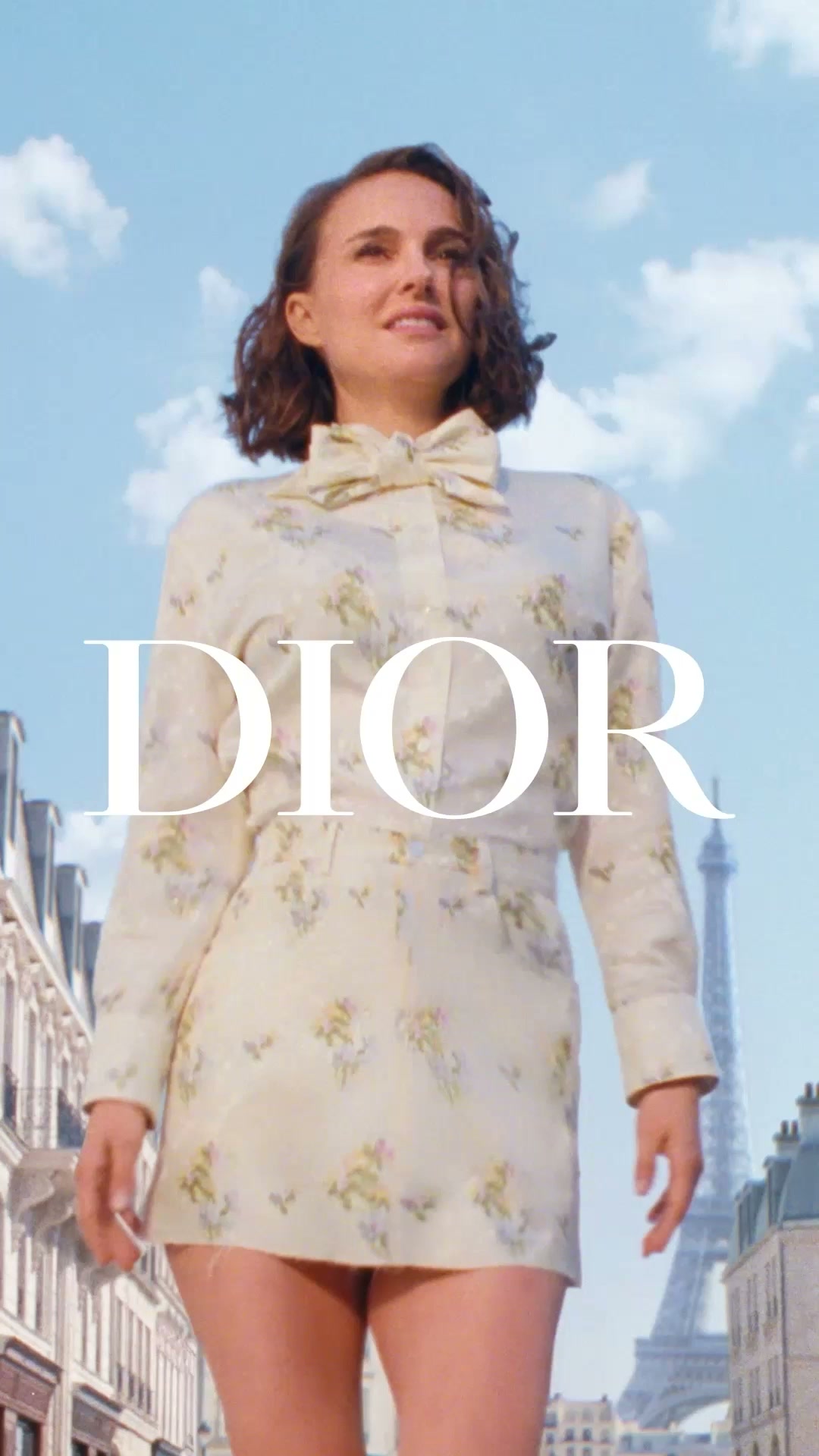 The ad features actress Natalie Portman in a floral dress and bow, standing in a Parisian street with the Eiffel Tower in the background. The Dior logo is prominently displayed in large white text across the center of the image. The text asks, "And you, what would you do for love?" The overall aesthetic is elegant and aspirational, evoking a sense of romance and high fashion.