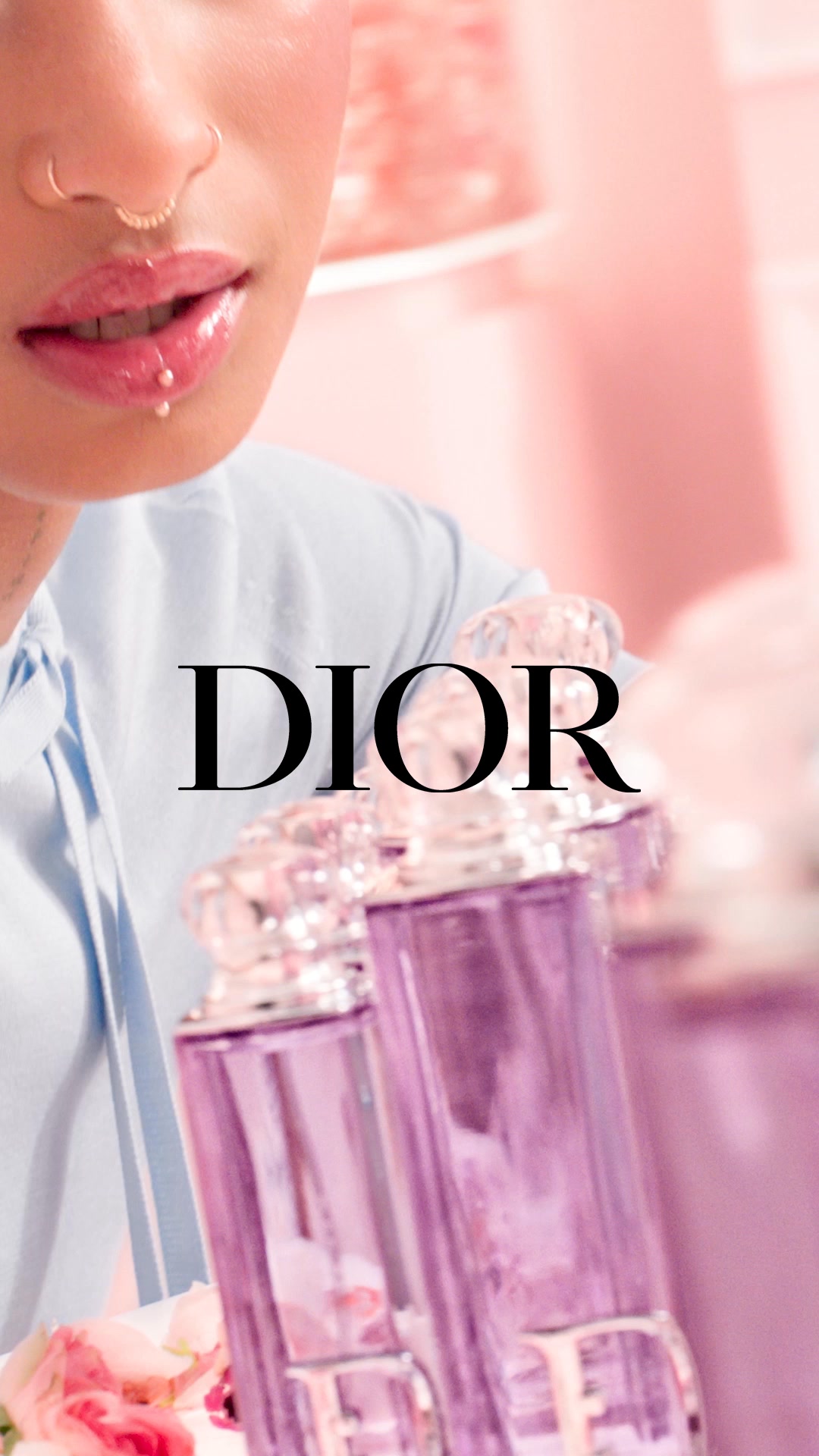 The image features a close-up of a person's face, focusing on their lips and nose, with delicate jewelry. They are wearing a light blue collared shirt. In the foreground, two ornate, purple glass perfume bottles are prominently displayed. The brand name "DIOR" is overlaid in large, black, serif font across the center of the image. The background is a soft, blurred pink, creating a dreamy and luxurious atmosphere. The text introduces the new Dior Addict fragrances, inspired by fruity floral notes, and mentions Willow Smith wearing Purple Glow.