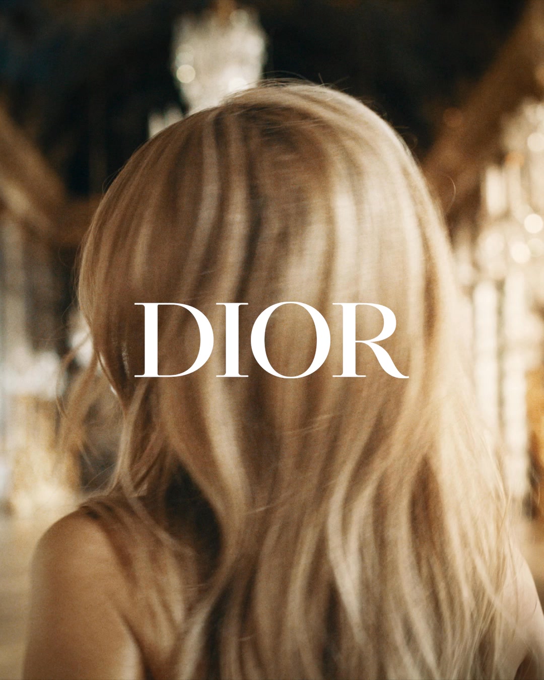 The image shows the back of a woman's head with long blonde hair. The word "DIOR" is prominently displayed in large white text across her hair. The background is blurred, suggesting an opulent indoor setting with chandeliers and ornate decor. The text overlay announces "J’ADORE, THE NEW INTENSE," positioning it as a bold claim about a new fragrance.
