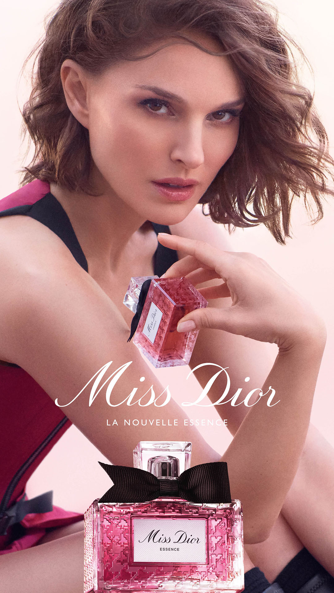 A close-up shot of actress Natalie Portman holding a bottle of Miss Dior perfume. The perfume bottle is pink and has a black bow. The text "Miss Dior" is written in elegant script across the lower half of the image, with "LA NOUVELLE ESSENCE" written below it. Another bottle of the perfume is visible at the bottom of the frame. The background is a soft, out-of-focus pink.