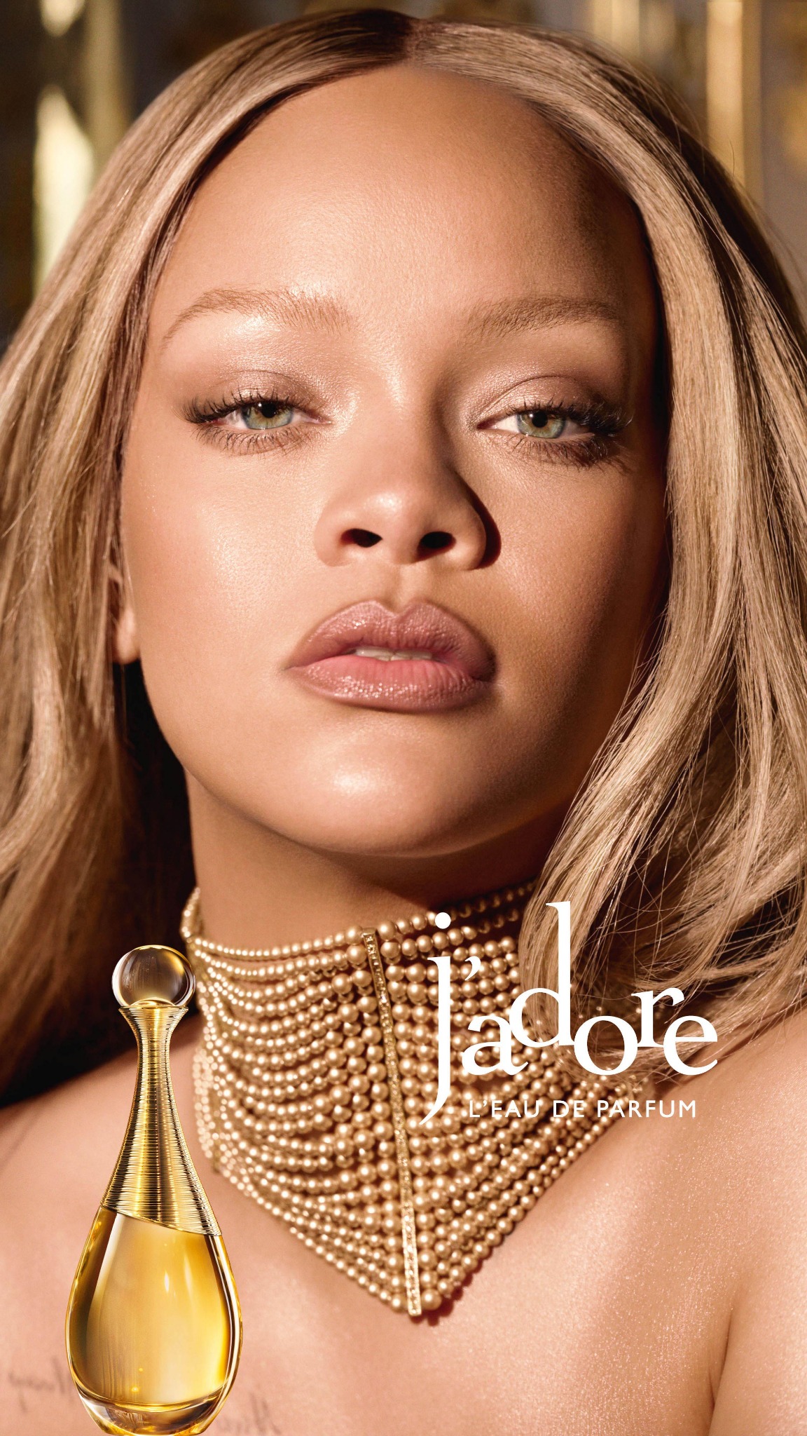 A close-up shot features a woman with blonde hair and striking green eyes, adorned with a pearl necklace. To the left, a bottle of J'adore Eau de Parfum is prominently displayed. White text overlays the image, reading "J'ADORE L'EAU DE PARFUM" and "Une icône rencontre un parfum de légende. Puissante, éblouissante et inspirante, Rihanna incarne la féminité triomphante et libre de #jadoredior." The overall aesthetic is luxurious and elegant, highlighting both the celebrity and the fragrance.