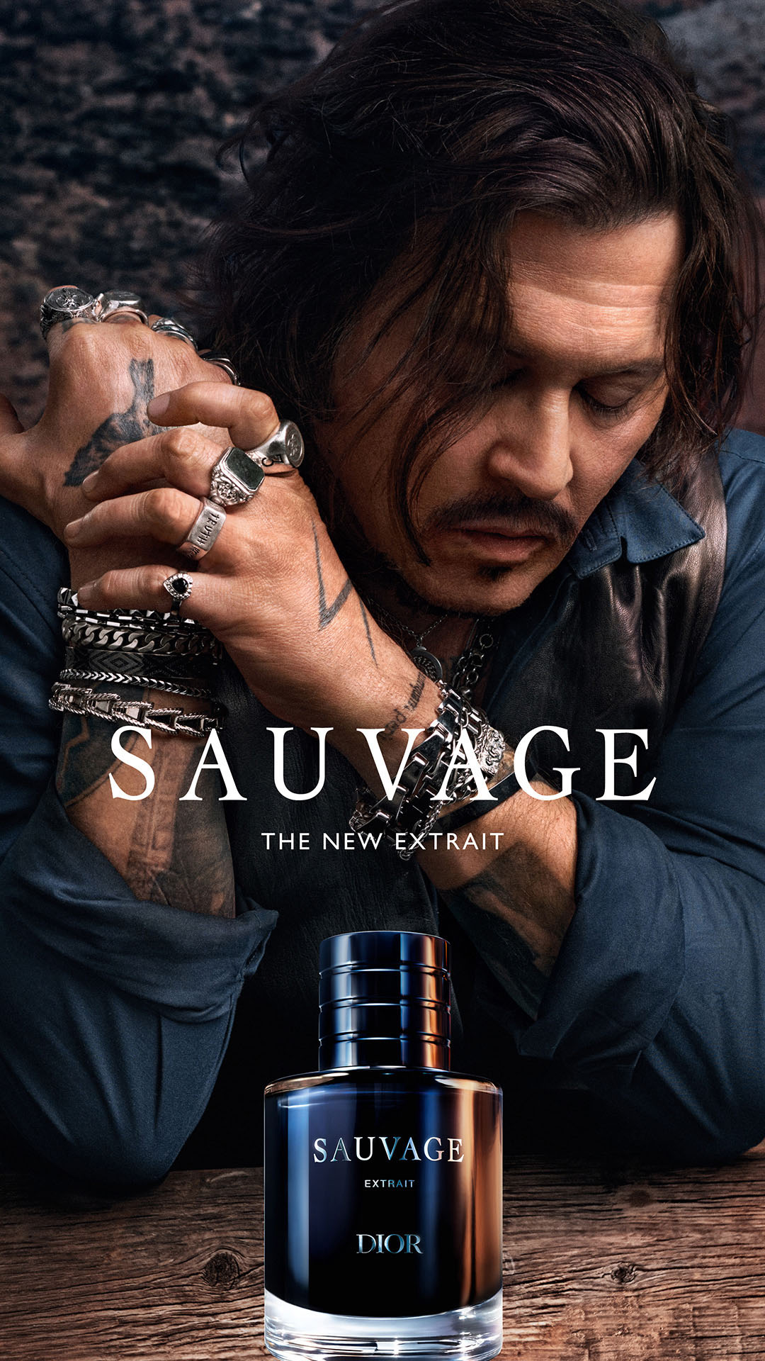 The image features a close-up of Johnny Depp, with his hands adorned with rings and bracelets, resting near his face. A bottle of Dior Sauvage Extrait perfume is prominently displayed in the foreground. The text "SAUVAGE, THE NEW EXTRAIT" is overlaid on the image, with smaller text below reading "THE NEW EXTRAIT". The overall aesthetic is dark, moody, and sophisticated, emphasizing the masculine and intense nature of the fragrance.