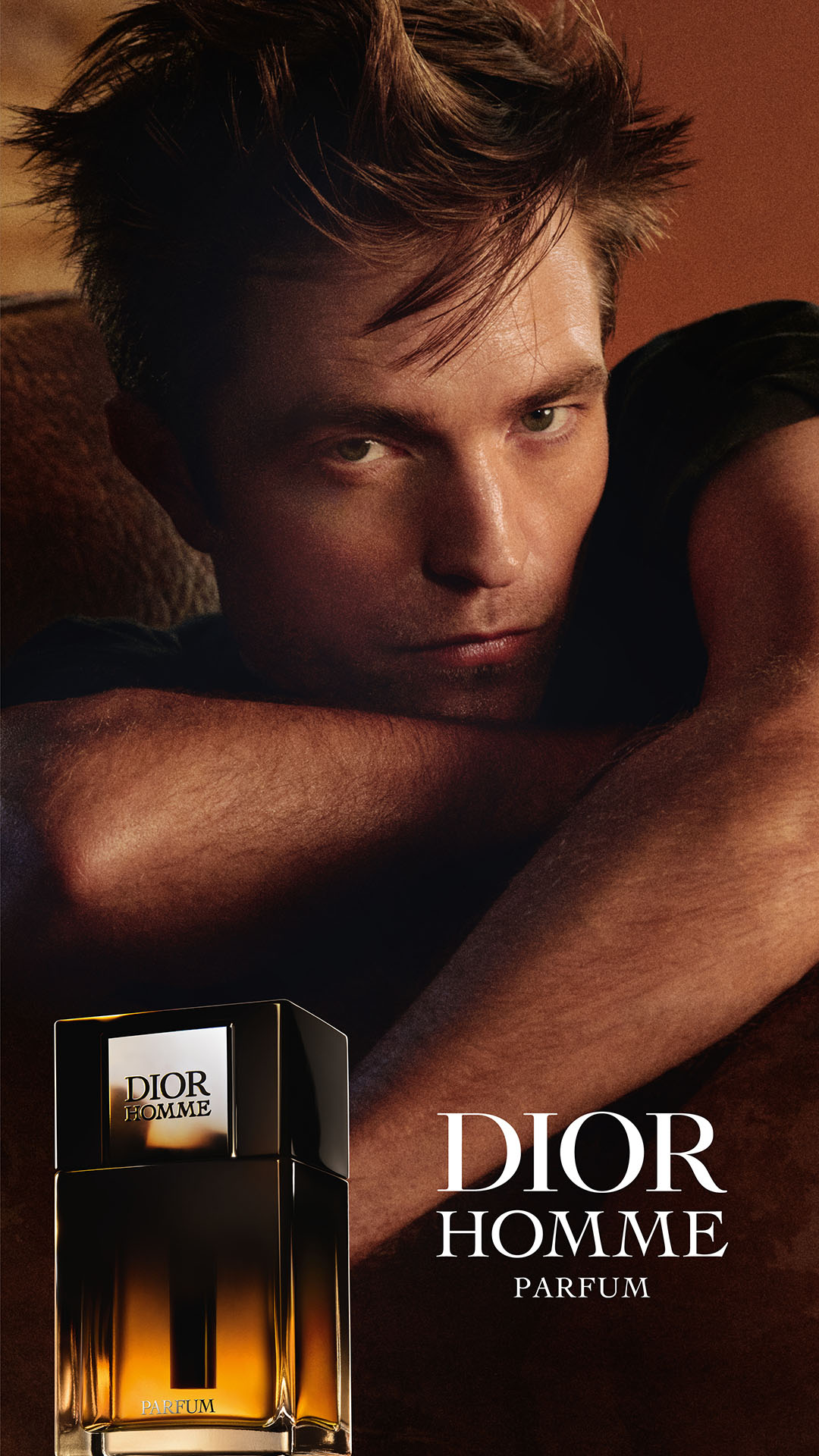 A close-up portrait of actor Robert Pattinson is featured, with his arms crossed in front of him. To the left, a bottle of Dior Homme Parfum is prominently displayed. The text "DIOR HOMME PARFUM" appears in large white font on the right side of the image, with "PARFUM" in smaller text below it. The overall aesthetic is dark and sophisticated, highlighting both the product and the celebrity endorser.
