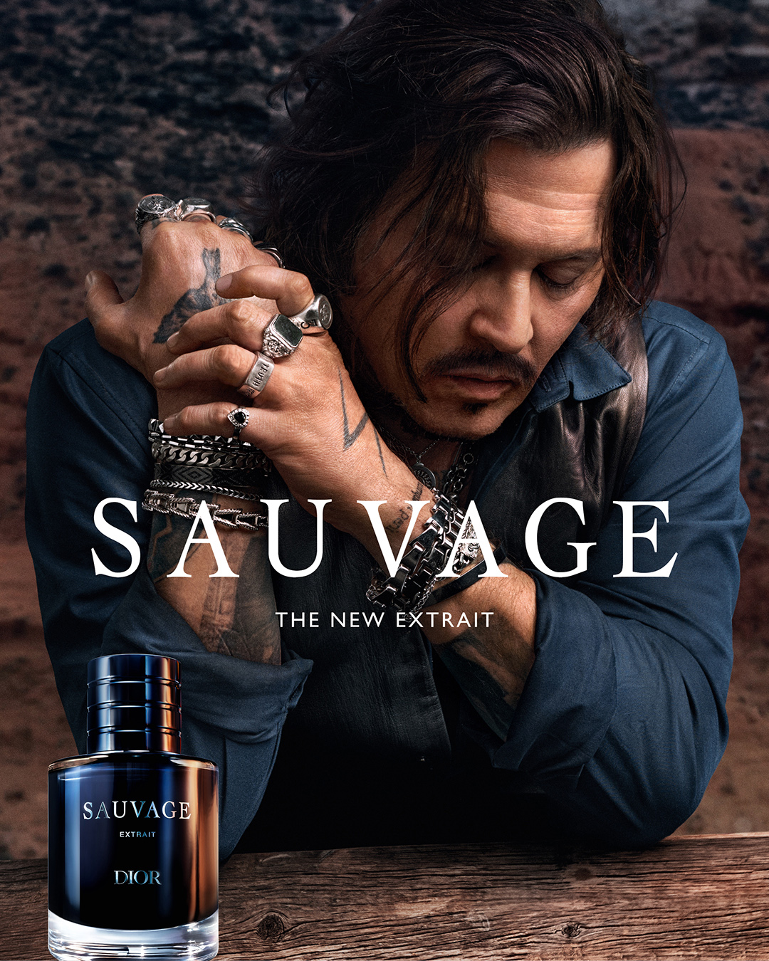 The creative features a close-up shot of Johnny Depp, with his hands clasped in front of his face, adorned with rings and bracelets. The text "SAUVAGE, THE NEW EXTRAIT" is prominently displayed in large white font across the image. Below this, smaller text reads "THE NEW EXTRAIT". A bottle of Dior Sauvage Extrait perfume is positioned in the lower-left corner. The background is a blurred, earthy landscape. The overall aesthetic is dark, intense, and sophisticated, highlighting the product and its celebrity endorser.