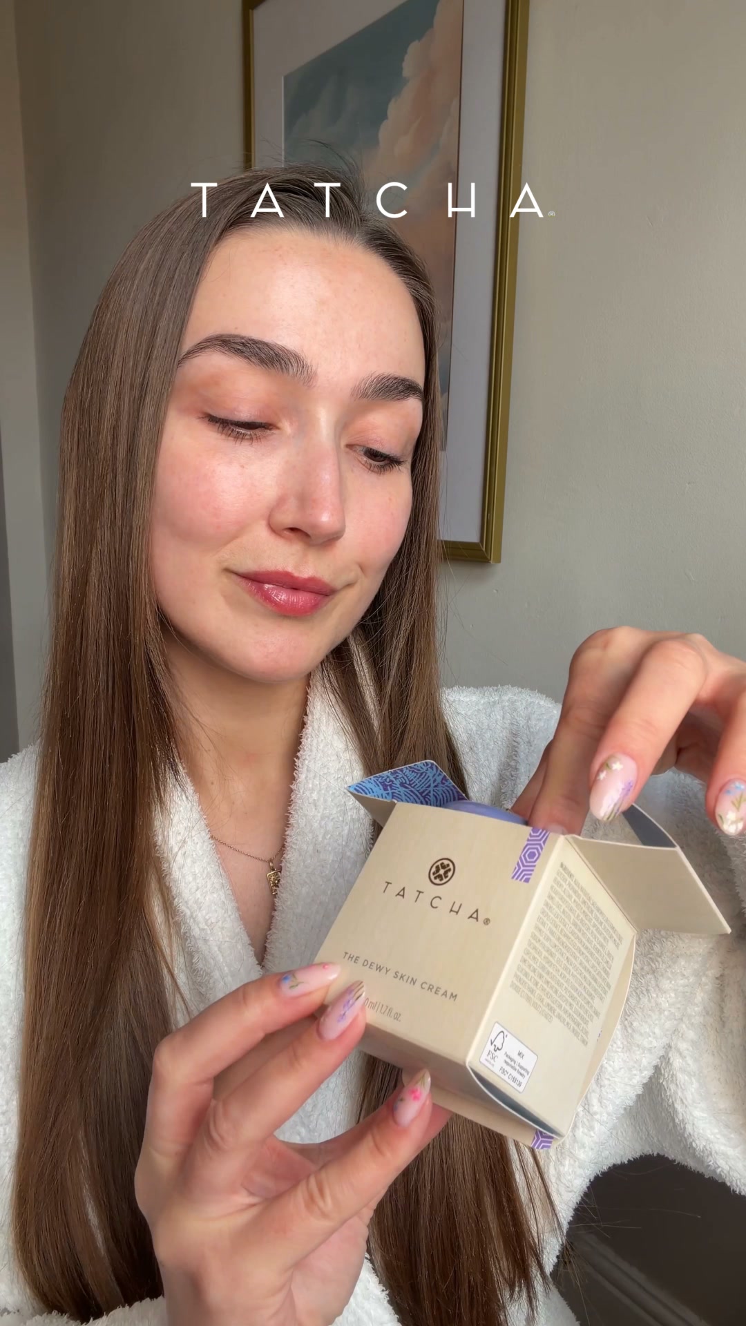 A woman wearing a white robe opens a Tatcha Dewy Skin Cream box. The Tatcha logo is visible on the box. Text overlays read "Scopri il best seller Dewy Skin Cream." and "Triplica l’idratazione della pelle all’instante." The woman has long brown hair and is looking down at the box she is opening. The background is a wall with a framed picture on it.