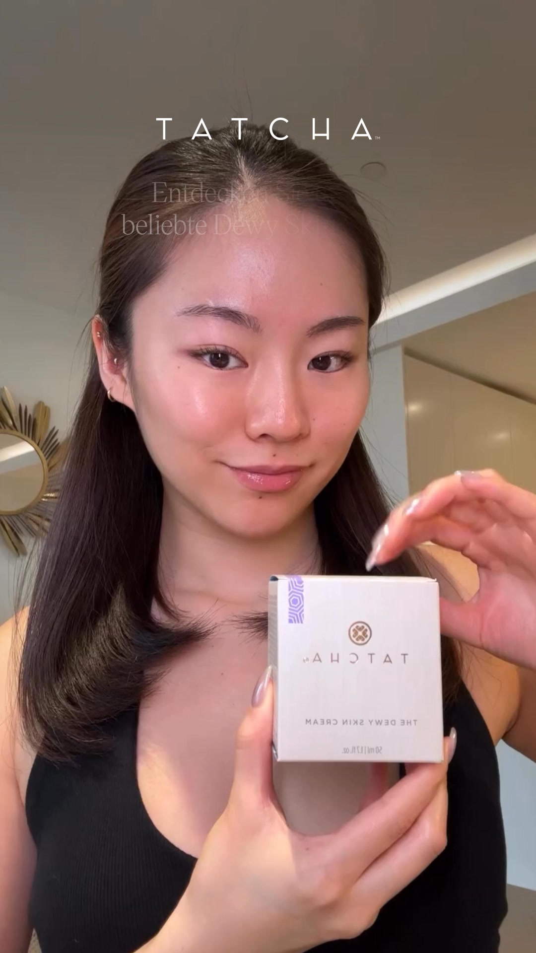 A young woman with clear skin smiles and holds up a small white box of Tatcha The Dewy Skin Cream. The brand logo and name are visible on the box. Text overlays the image, stating "Erhöht augenblicklich die Hautfeuchtigkeit um das Dreifache. Testen Sie The Dewy Skin Cream." The background is a blurred indoor setting.