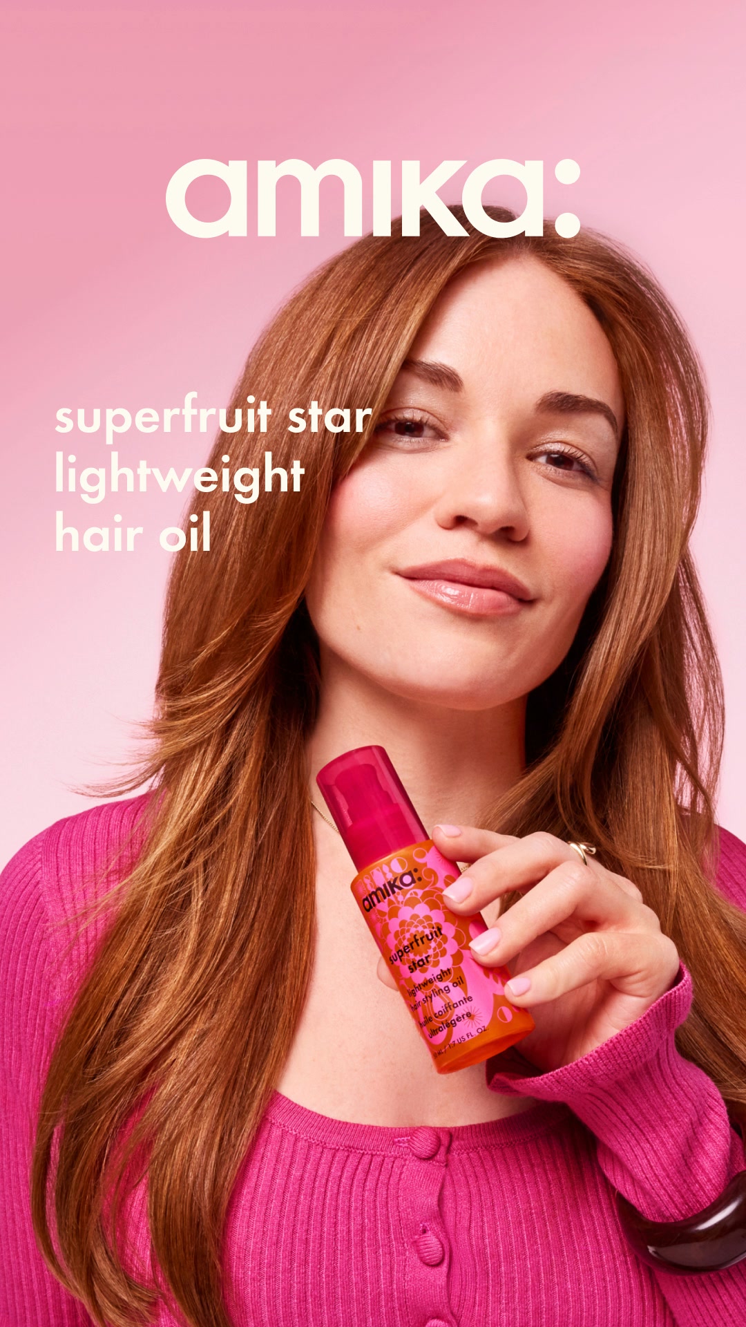 A woman with long, auburn hair is holding a bright pink bottle of Amika Superfruit Star Lightweight Hair Oil. She is wearing a pink ribbed sweater and looking towards the camera with a slight smile. White text overlays the image, reading "amika:" at the top and "superfruit star lightweight hair oil" on the left side. The background is a soft pink gradient. The ad focuses on the product and its benefits for hair.