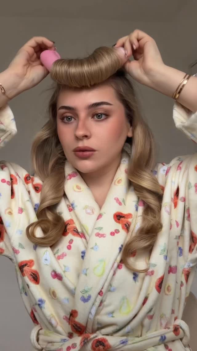 A woman with blonde hair is shown in a robe, using pink rollers to style her hair. She is demonstrating a hair styling technique. On-screen text reads 'how to fake a blowout at home⭐️ these 3 products by @amika are a must in my routine! they keep my hair looking healthy, shiny & silky 👱🏼‍♀️✨'. The video appears to be a tutorial on achieving a blowout look without heat styling.