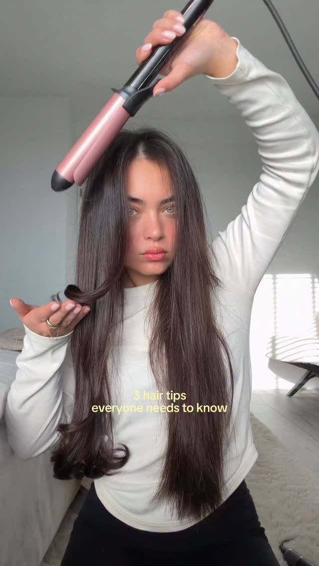 A young woman with long, dark hair is shown from the chest up, holding a curling iron above her head. She is demonstrating a hair styling technique, with a strand of her hair curled and held out in her left hand. On-screen text reads "3 hair tips everyone needs to know." The background is a softly lit room with a window casting striped shadows.