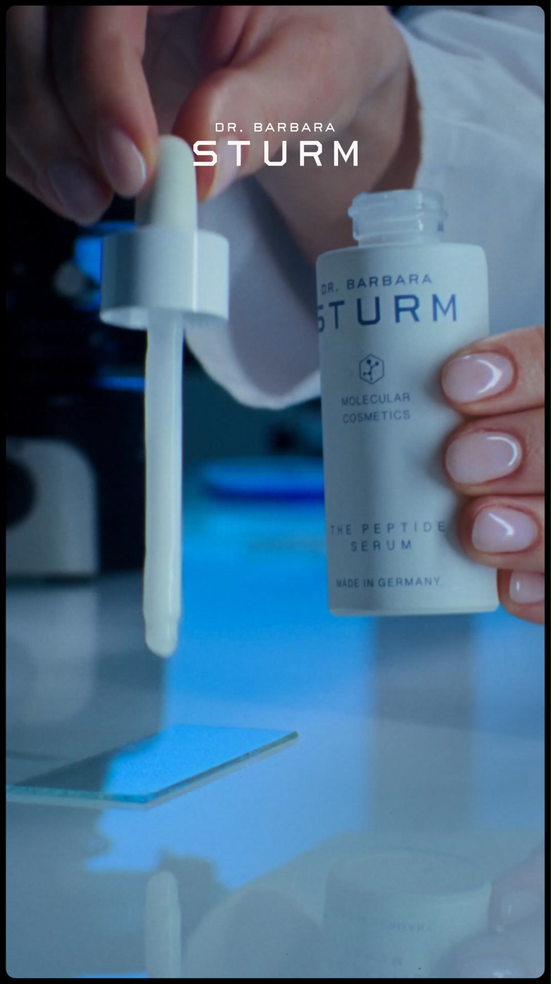 A close-up shot shows a hand holding a small white bottle of Dr. Barbara Sturm The Peptide Serum. The dropper is being held over a glass slide, with a drop of serum suspended from the tip. The bottle's label is visible, stating "THE PEPTIDE SERUM" and "MADE IN GERMANY." The text "DR. BARBARA STURM" is prominently displayed at the top of the bottle and also in the background. The overall scene suggests a laboratory or clinical setting, emphasizing the scientific formulation of the product.