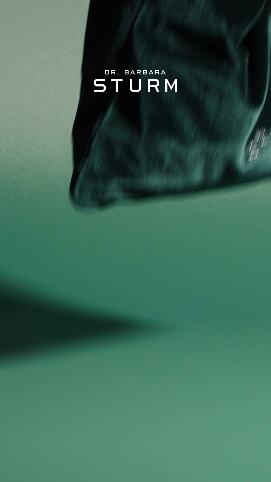 The creative features a close-up, slightly blurred shot of a dark green, textured fabric, possibly a bag or garment. The brand name "DR. BARBARA STURM" is prominently displayed in white, sans-serif text across the upper portion of the fabric. The background is a solid, muted green, with a soft shadow cast from the upper object onto the lower half of the frame. The text overlay explains the Dr. Barbara Sturm Winter Kit, a 7-step routine for soothing and nourishing skin, and mentions a limited-edition velvet-green bag.