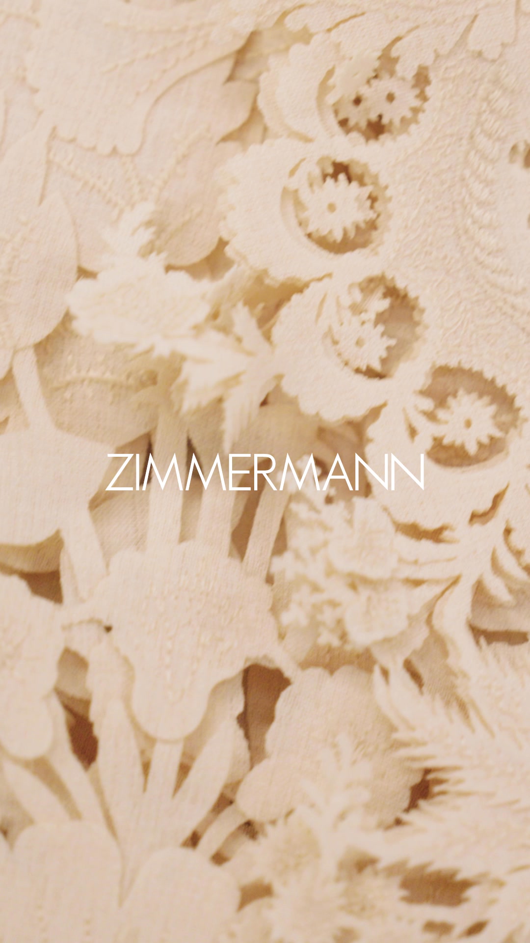 The creative is a close-up, textured image of cream-colored fabric with intricate floral cutouts. The brand name "ZIMMERMANN" is displayed in white, sans-serif capital letters across the center of the image. The fabric appears delicate and detailed, suggesting a high-end garment. The overall aesthetic is elegant and focused on the material and branding.