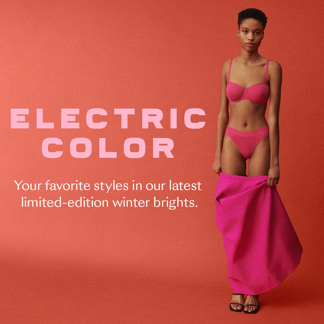 A model stands against a coral-colored background, wearing a pink bra and underwear. She is holding a large piece of pink fabric. The words "ELECTRIC COLOR" are displayed in large, light pink sans-serif font on the left side of the image. Below the headline, the text reads "Your favorite styles in our latest limited-edition winter brights." The overall aesthetic is clean and modern, highlighting the vibrant color of the apparel.