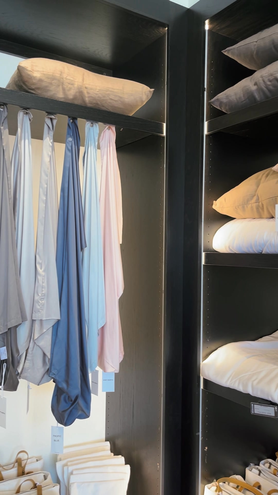 The creative displays a well-organized closet with shelves and a hanging rack. On the rack, several pieces of fabric in muted colors like grey, blue, and pink are neatly hung. The shelves above and to the side are stacked with pillows and bedding in various neutral tones. The overall impression is one of calm, order, and comfort, showcasing the brand's focus on home textiles and linens.