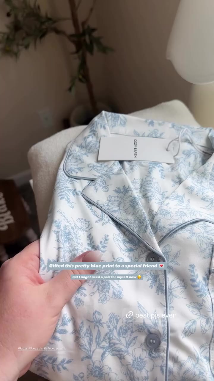 A close-up shot of a light blue and white floral print pajama set from Cozy Earth. The pajamas are folded and a hand is holding them up, showing the fabric and buttons. Text overlays read "Gifted this pretty blue print to a special friend" and "But I might need a pair for myself now 🥺". Another overlay at the bottom says "@best pjs ever" and hashtags "#Cozy #CozyEarth @cozyearth".