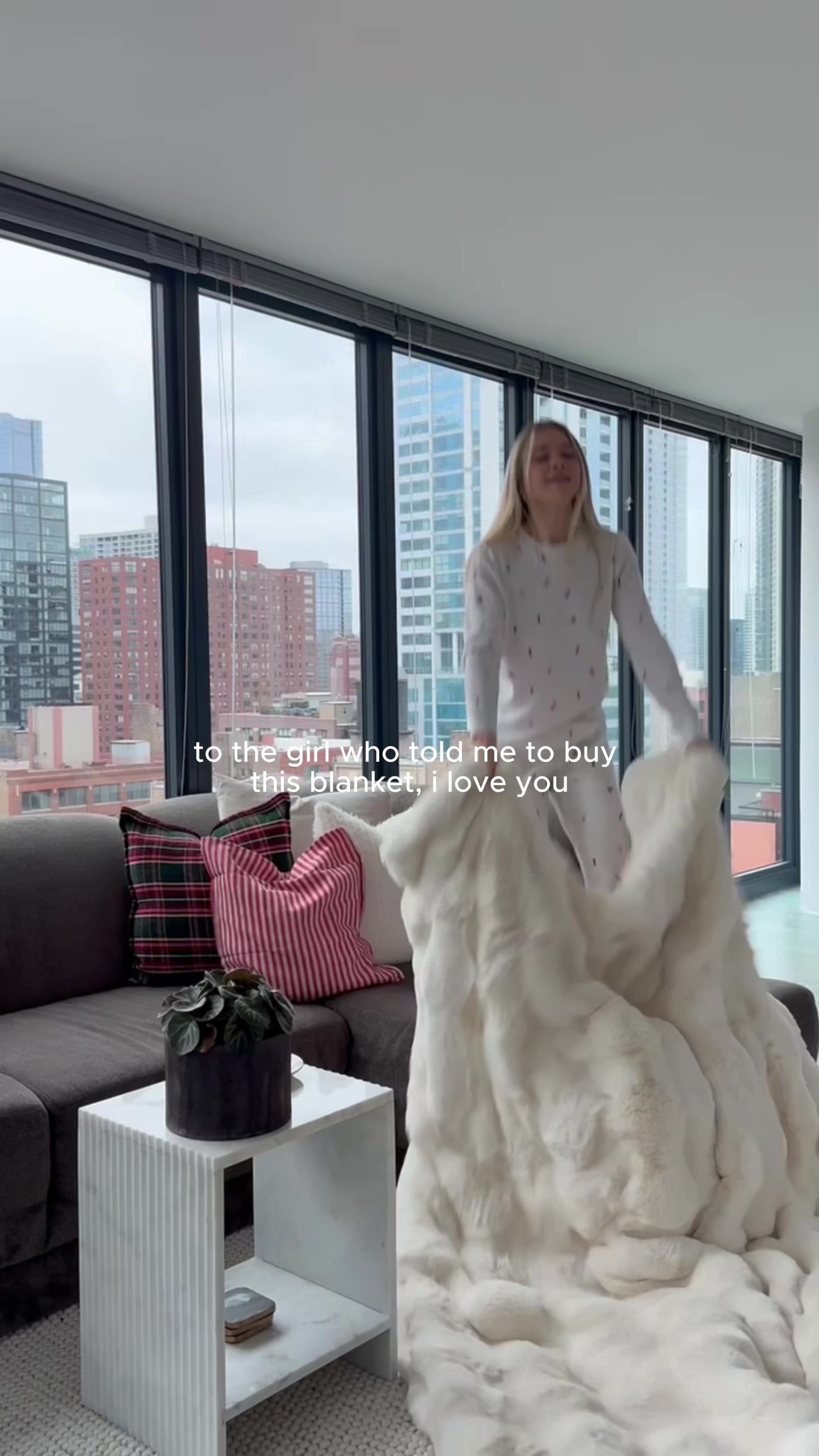 A woman is shown in a living room, holding up a large, fluffy white blanket. She is wearing a light-colored sweater with a subtle pattern. The text overlay reads "to the girl who told me to buy this blanket, i love you". The background features large windows with a city skyline visible outside, a sofa with decorative pillows, and a small side table with a plant. The overall scene conveys a sense of comfort and appreciation for a recommended product.