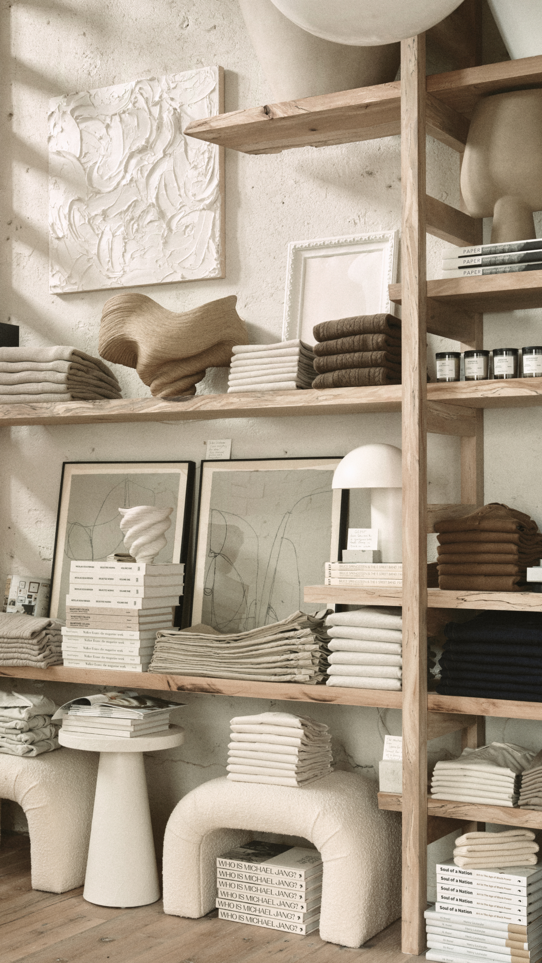 A well-lit, neutral-toned image displays a retail store's shelving unit filled with folded clothing, books, and decorative items. The shelves are made of light-colored wood, and the background is a textured, off-white wall. The overall aesthetic is minimalist and organized, showcasing a curated selection of home goods and apparel. There is no text visible on the creative itself.