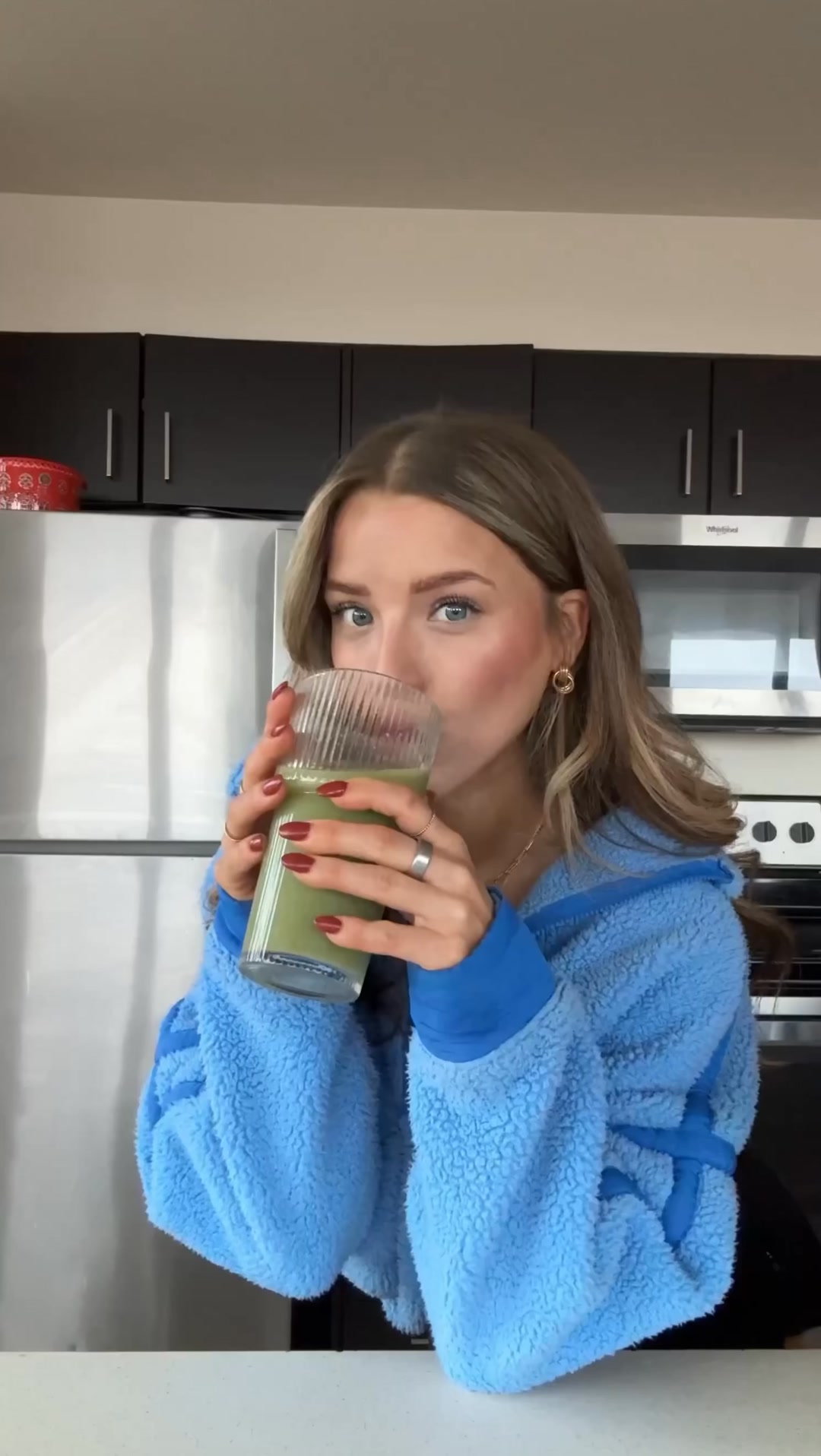 A young woman with blonde hair, wearing a blue fuzzy robe, is drinking a green beverage from a clear glass. She is in a kitchen setting with stainless steel appliances and dark cabinets visible in the background. Text overlaying the image reads: "A daily wellness routine thats actually worth doing."