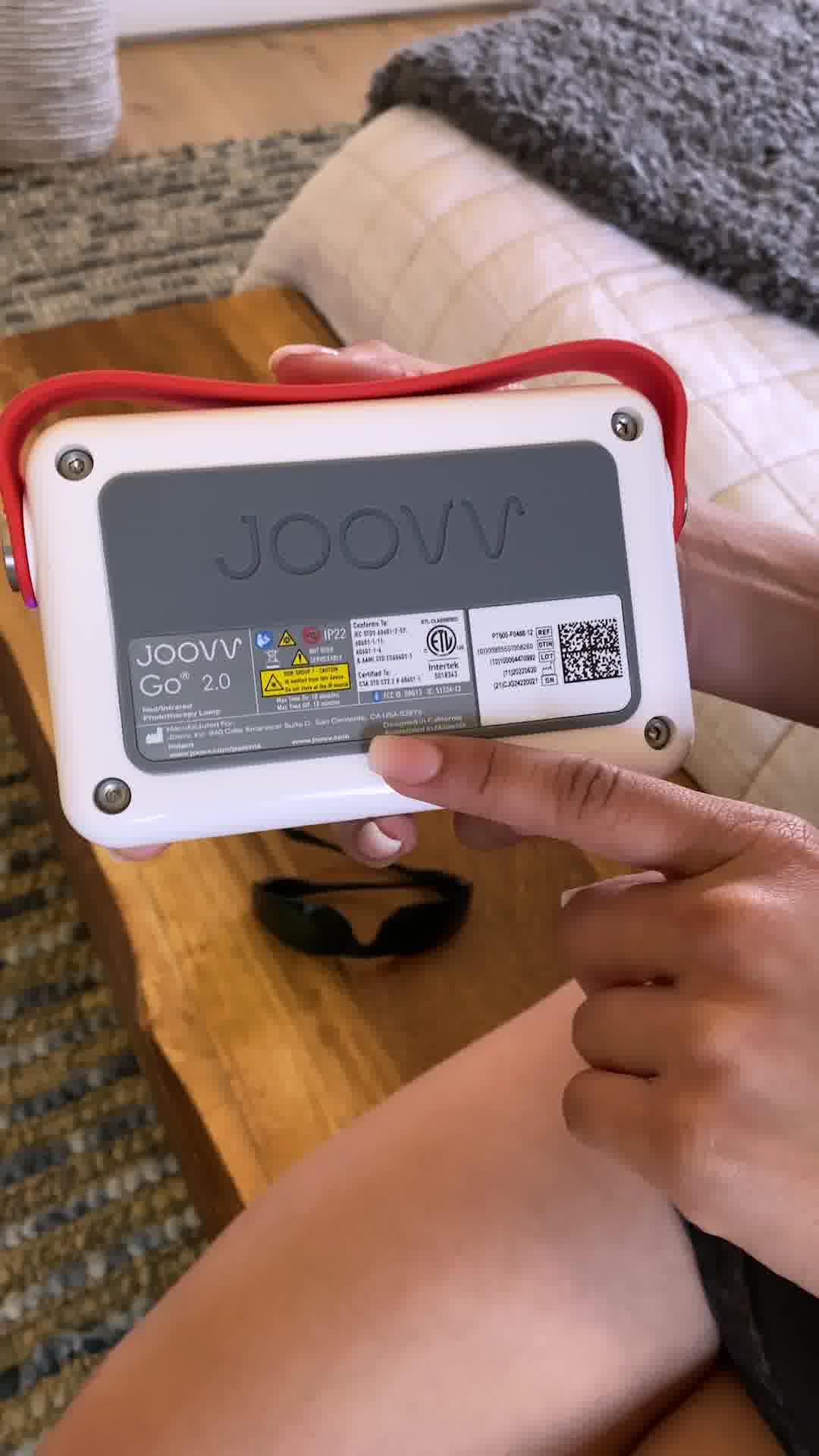 A person's hand is shown holding a white and gray Joovv Go 2.0 device with a red handle. The device has various labels and certifications printed on its back. In the foreground, another hand is pointing to the device. Below the device, a pair of black protective glasses rests on a wooden table. The background shows a blurred view of a couch and a rug.