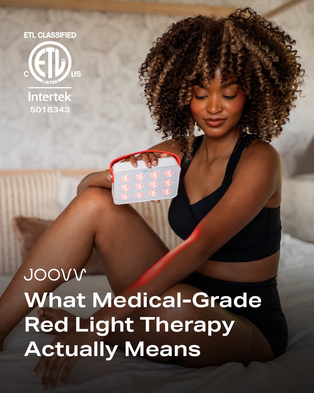 A woman with curly hair sits on a bed, holding a white and red portable red light therapy device. The device has a grid of red lights, and a red beam of light is visible on her arm. Text overlays on the image read "ETL CLASSIFIED", "Intertek 5018343", "JOOVV", and "What Medical-Grade Red Light Therapy Actually Means". The overall image is a lifestyle shot with informational text about red light therapy.