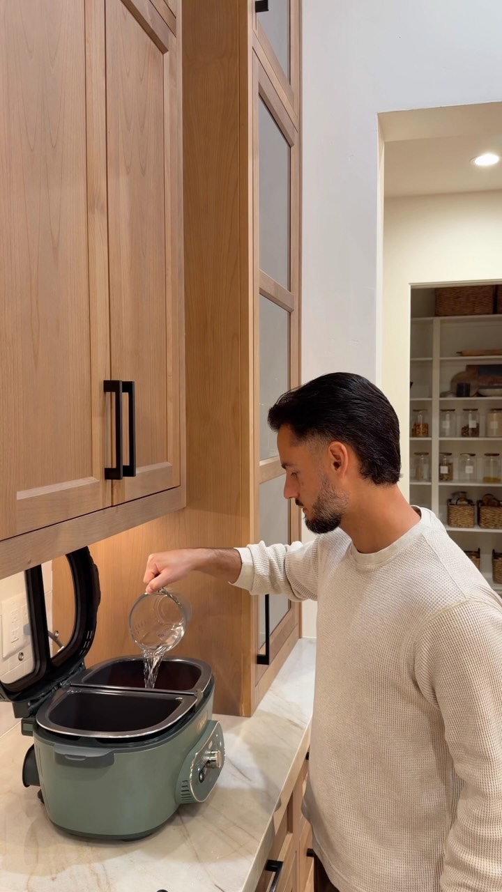 A man in a cream-colored waffle-knit sweater is in a modern kitchen, pouring water from a glass measuring cup into a dual-compartment green slow cooker. The cabinets are light wood with black handles. The hook text overlays the image, describing the relatable struggle of weeknight cooking and the desire to be present with family. The man is identified as a #NinjaPartner in the ad copy.