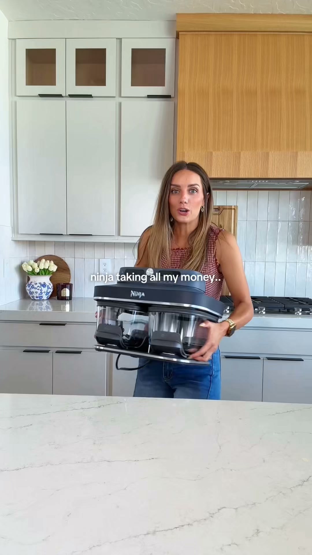 A woman stands in a modern kitchen holding a Ninja Crispi DualZone appliance. She is wearing a red and white checkered top and blue jeans. On-screen text reads "Ninja taking all my money..". The kitchen has white cabinets, wood accents, and a white marble countertop. The woman is looking towards the camera with a slightly exasperated expression, implying she has spent a lot on Ninja products but finds them worthwhile.