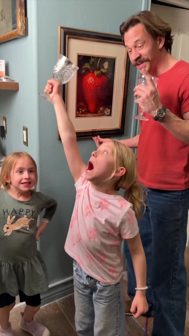A man and two young girls are in a room. The girl in the middle is holding up a large wine glass filled with a clear liquid, with her mouth open in excitement. The man on the right is smiling and holding a similar glass. The girl on the left is also smiling and looking towards the camera. A framed picture of a large strawberry hangs on the wall behind them. Text overlays the image: "Slushi on Tap. Every kitchen needs this!" and hashtags related to Ninja Kitchen and Slushi.
