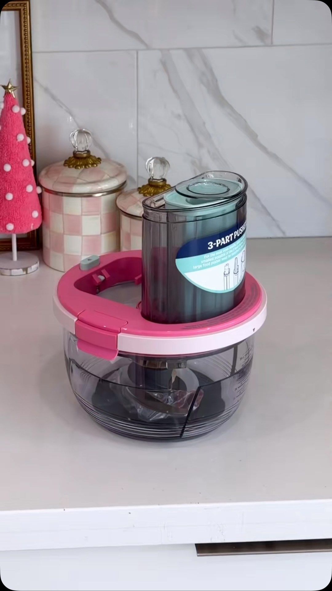 A pink and white kitchen appliance, a food chopper, is shown on a white countertop. The appliance has a clear bowl and a pink lid. A small, decorative pink Christmas tree with white pom-poms is visible to the left of the appliance. The text "FOR THE PINK GIRLIES 💝💖😍" is overlaid on the image in a playful font.