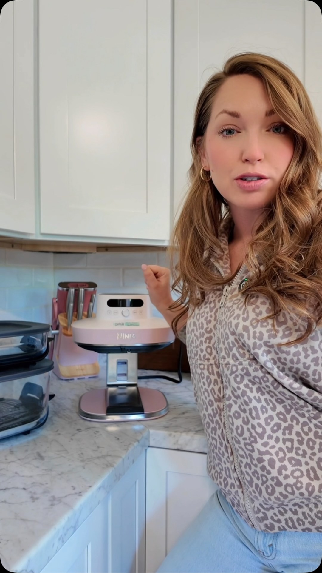 A woman with long, wavy blonde hair is standing in a kitchen, gesturing towards a pink Ninja air fryer on the counter. She is wearing a leopard-print zip-up hoodie and light blue jeans. The text overlay reads, "Your air fryer could NEVER look this cute on the counter 😭🩷". The kitchen has white cabinets and marble countertops. Other kitchen appliances are visible in the background.
