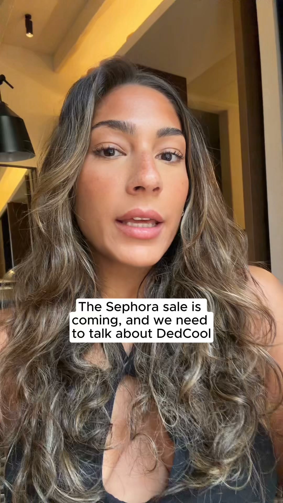 A close-up shot of a woman with long, wavy brown hair and light makeup. She is looking slightly off-camera with her mouth slightly open. White text is overlaid on the bottom half of the image, reading: "The Sephora sale is coming, and we need to talk about DedCool". The background is blurred, showing a room with warm lighting and a dark lamp on the left.
