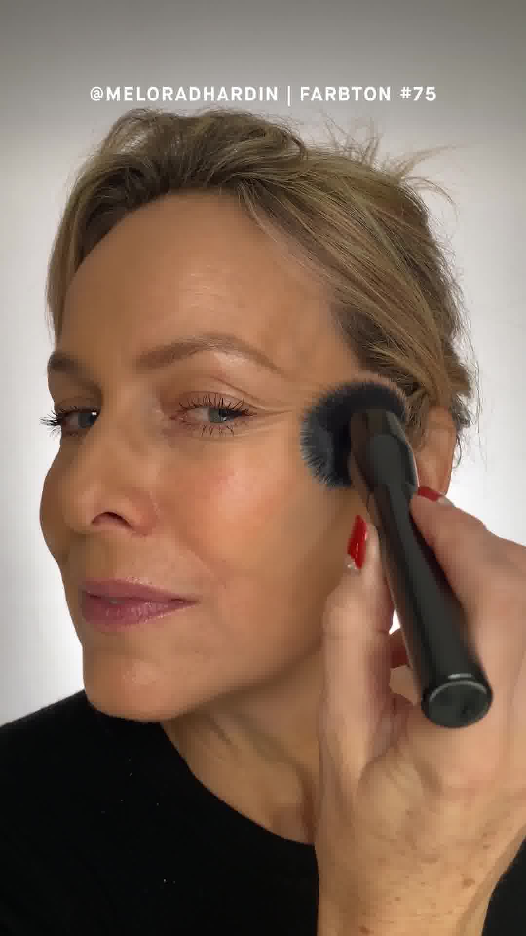 A close-up, profile view of a woman's face as a makeup brush is used to apply product to her cheek. The woman has blonde hair pulled back and is looking towards the left of the frame. Text overlayed on the image reads "@MELORADHARDIN | FARBTON #75". The copy states, "As my skin is aging now, I am VERY picky about what I put on my face. This foundation is magic in a bottle! It instantly makes me look 10 years younger." - Alex.