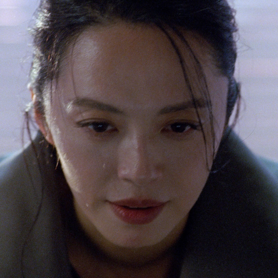 A close-up, low-angle shot focuses on the face of a woman with dark hair pulled back, looking downwards. Her skin appears dewy, with a single drop of sweat visible on her cheek. She is wearing a dark jacket or coat. The background is softly blurred with a cool, muted tone. The image is a still from a campaign, featuring a model.